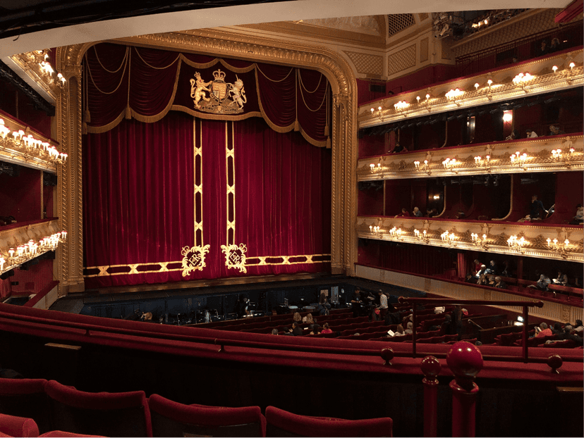 Interior of an ornate theater with a red velvet curtain and gold accents, surrounded by multiple tiers of seating.